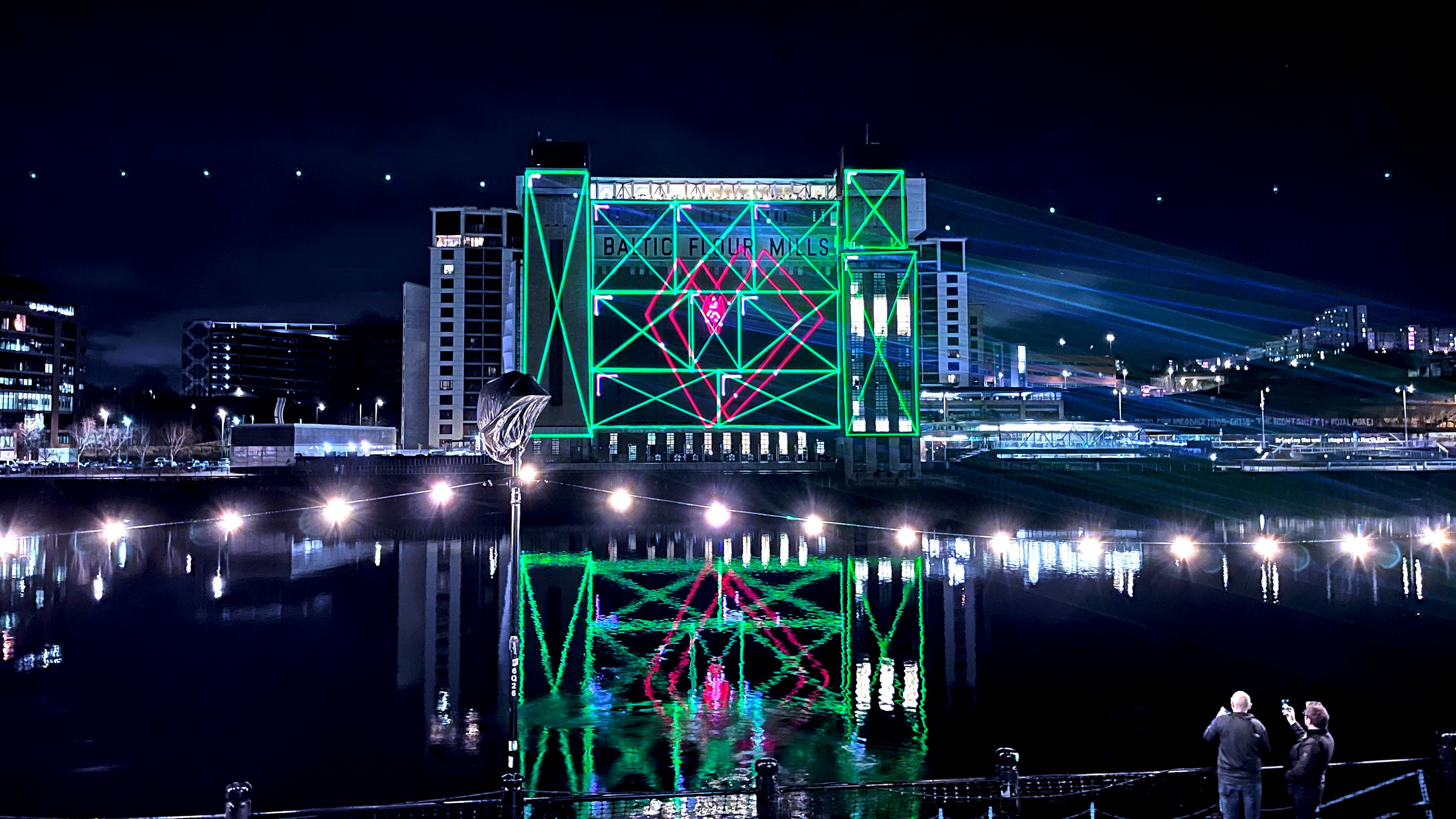 Green and magenta laser graphics mapped onto the BALTIC building in Newcastle, reflected in the River Tyne